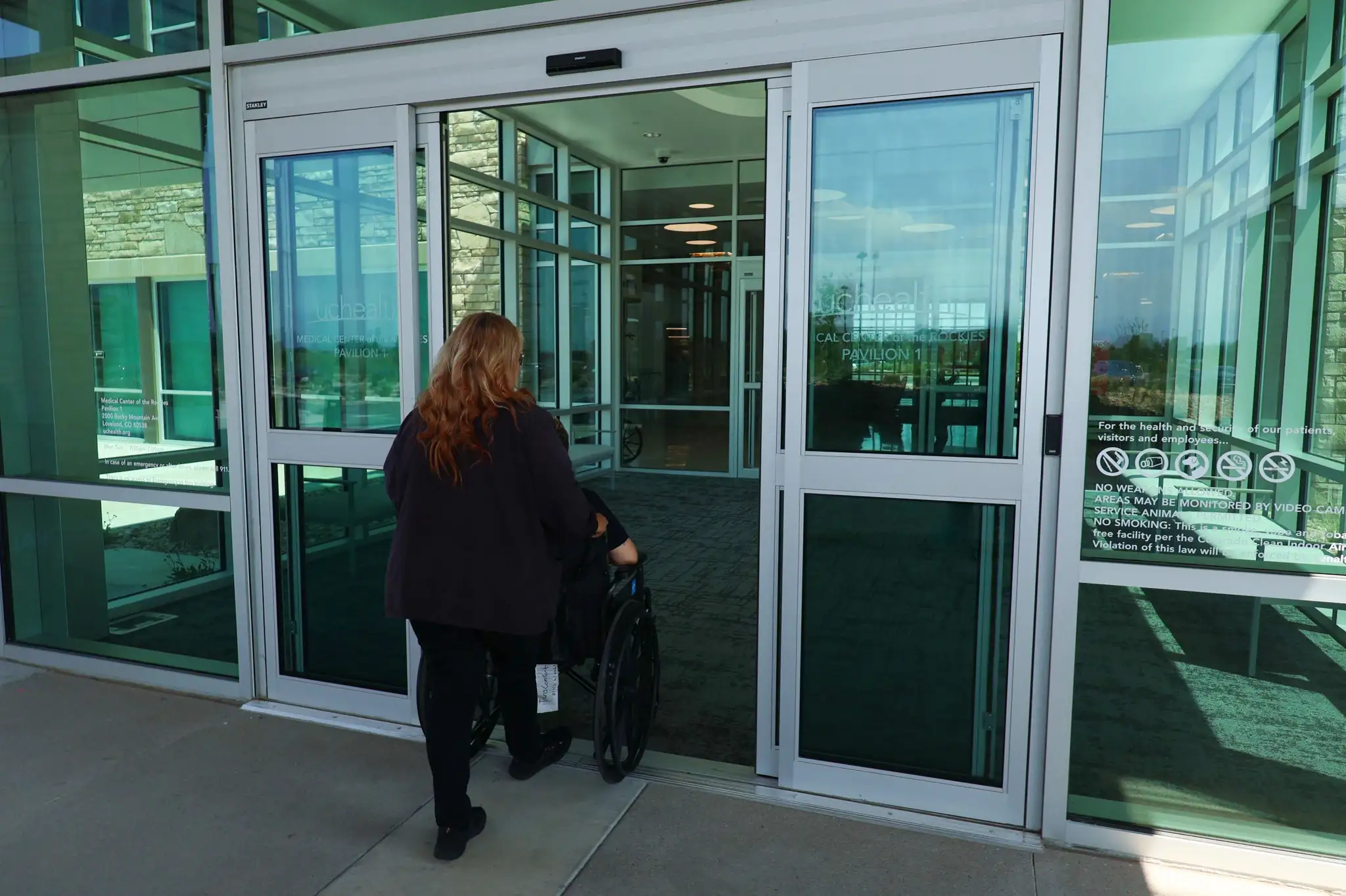 A TransComfort attendant pushing a seated passenger through the automatic doors into a hospital pavilion.